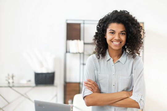 Young Pleasant Brunette African American Mixed-race Woman With Afro Hairstyle Wearing A Casual Shirt, Standing With The Arms Folded. Smiling And Posing Against Blurred Home Office Interior Background