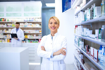 Portrait of smiling female blonde pharmacist standing in pharmacy shop or drugstore with her arms...