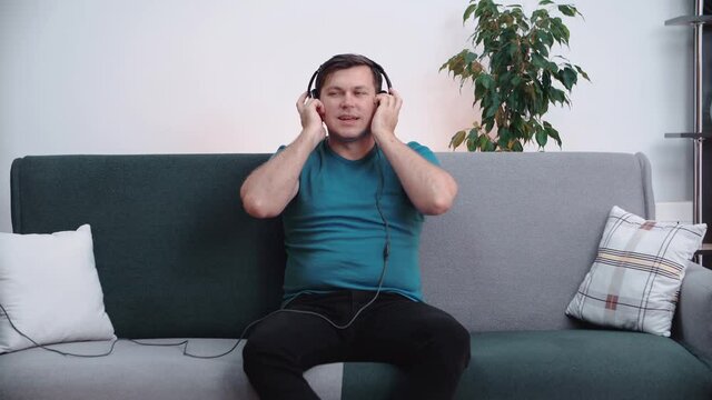 A man sits on the couch wearing headphones on his head, quietly listening to music.