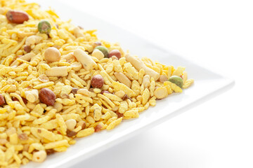 Delicious Khatta Meetha in a white ceramic square plate, made with peanuts, sugar, raisins, and besan sev. Pile of Indian spicy snacks (Namkeen), under backlight, side view