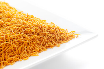 Spicy Aloo Bhujia in a white ceramic square plate, made with Red chili, Potatoes, and Chickpeas. Pile of Indian spicy snacks (Namkeen), under backlight, side view, against the white background.