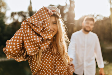 Fototapeta premium Blue-eyed smiling woman puts on beige cap and looks into camera, holding her boyfriend's hand. Girl in brown dress covers her face with her hand