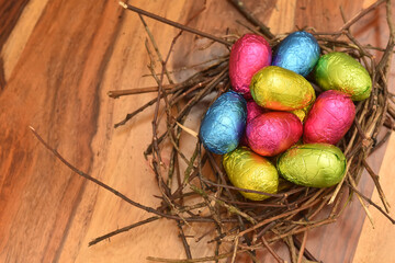 Foil wrapped colourful easter eggs in pink, green, blue and yelow in a natural nest made of sticks and twigs, against a multi grain brown wooden background.