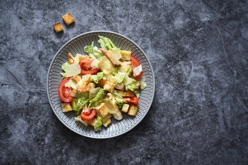 Classic Caesar salad with chicken, tomato and sauce in a plate on a dark concrete background, top view.