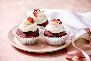 Homemade Red velvet cupcakes with whipped cream on pink ceramic plate, white napkin with ribbon over pink texture background.