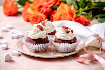 Homemade Red velvet cupcakes with whipped cream on pink ceramic plate, white napkin with ribbon, roses flowers, wooden hearts over pink texture background. Valentines day dessert.