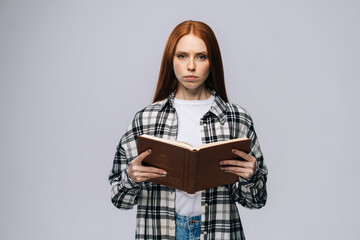Fototapeta premium Portrait of serious young woman college student holding opened books and looking at camera on gray background. Pretty redhead lady model wearing casual clothes emotionally showing facial expressions