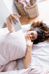 Smartphone in hand of woman taking selfie on blurred background on bed