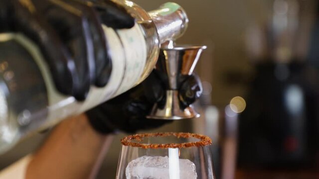 Bartender preparing a cocktail on a bar counter. By pouring alcoholic beverage from a bottle into a small metal measuring cup and then into a glass cup with ice inside.