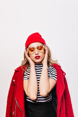 Fototapeta na wymiar Curly girl with red lips in hat and glasses massages her head. Blonde in bright jacket and striped T-shirt posing on white background