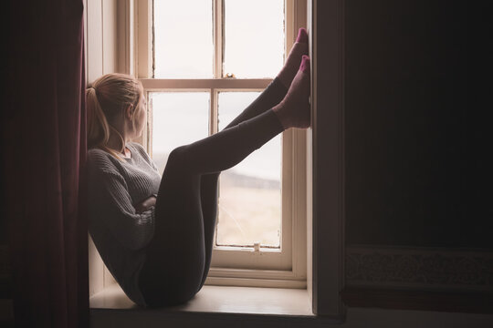 A young woman stuck indoors sits on a window sill, gazing outside and longing to be free.
