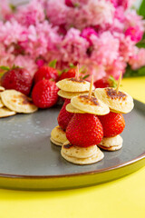 Mini pancakes with strawberries strung on toothpicks, a kind of breakfast. In the background are pink flowers.