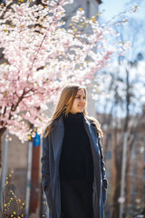 Sakura branches with flowers on a tree on the city streets. Happy woman girl in a gray palette walks along an alley with blooming sakura. Gorgeous fancy girl outdoors. Sakura tree blooming.