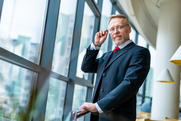 Hansome mature grey haired man touches spectacles with hands. Male wears dark suit and red tie. Big panoramic windows with city view blurred background.