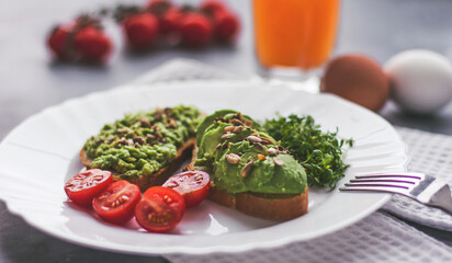 A vegan avocado and seed sandwich sits on a white plate with tomatoes on a gray background