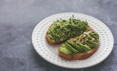 A vegan sandwich with avocado and seeds sits on a white plate on a gray background