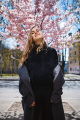 Young slender female model with long wavy hair and, dressed in a gray coat, sneakers, spinning on the street. Spring blossom trees woman girl laughs and runs to rejoice