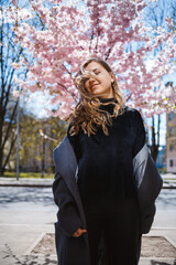 Fototapeta premium Young slender female model with long wavy hair and, dressed in a gray coat, sneakers, spinning on the street. Spring blossom trees woman girl laughs and runs to rejoice