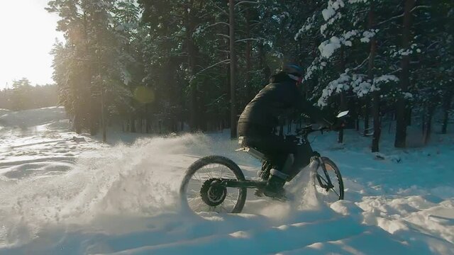Cyclist in Black Clothes, Helmet and Snow Glasses Riding on Electric Bike in the Snow in Winter Forest. E-Bike Riding and Drifting at Cold Sunny Day - Super Slow Motion