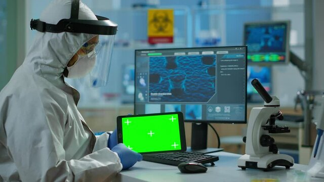 Doctor with coverall working at tablet with green screen in modern equipped lab. Team of microbiologists doing vaccine research writing on device with chroma key, isolated, mockup display.