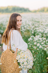 Beautiful girl with daisies bouquet, back view. Tranquil summer in countryside. Stylish young woman in white vintage dress and hat waling on field of daisies.