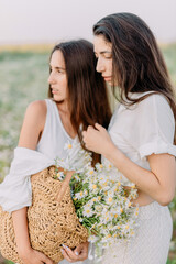 Two young unusual brunette girls in white clothes stand and pose in the middle of a blooming meadow of chamomile with a bouquet of wildflowers. Enjoy the summer and nature. Fashionable look