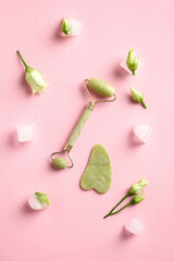 Jade stone face roller and gua sha on pink background with ice cubes and flowers. Skincare concept. Flat lay, top view.