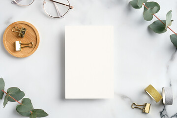 Blank white greeting card with feminine accessories, office supplies and eucalyptus leaves on marble desk table. Flat lay, top view, copy space.