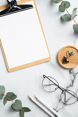 Elegant feminine workspace with clipboard, glasses, office supplies, eucalyptus branches on marble background. Home office desk table. Flat lay, top view.