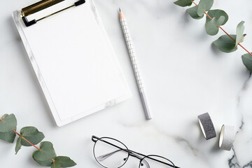 Minimal style feminine workspace with clipboard, glasses, ribbon and eucalyptus leaves on marble background. Elegant home office desk table.