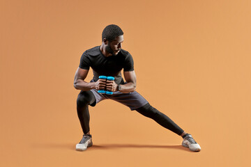 Fototapeta na wymiar african american fitness sportsman is doing exercise squatting with dumbbells, isolated on brown studio background
