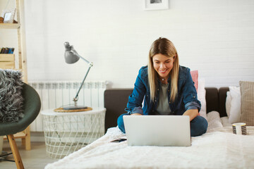Young woman sitting on bed in bedroom and using laptop