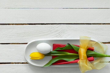 Egg and yellow tulip decorative aranged as mimal bouquet with bow on plate with white wooden background. Creative Easter idea and concept. Flat lay.