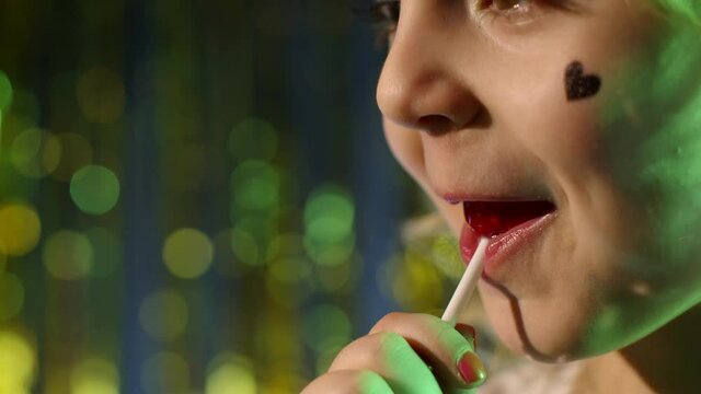 Close-up shot of stylish teenager caucasian kid girl lips licking lollipop with tongue, slow motion 4k footage. Child in night club backlit with blue neon light. Futuristic style, sensual look clubber