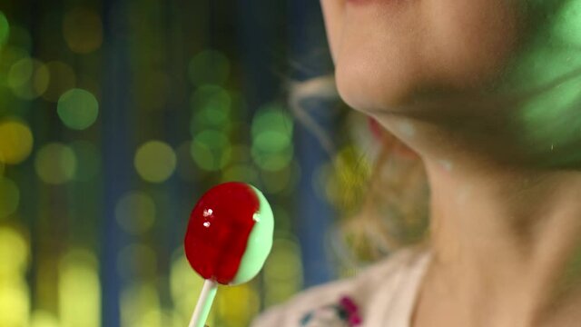 Close-up shot of futuristic hipster teen girl licking lollipop candy in slow motion at disco party cyberpunk club. Funny caucasian child kid in neon lights on illuminated wall. Sensual look clubber