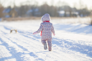 Happy baby girl in overalls running on white snow covered country road in sunny winter day. Back...
