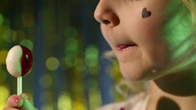 Close-up shot of stylish teenager caucasian kid girl lips licking lollipop with tongue, slow motion 4k footage. Child in night club backlit with blue neon light. Futuristic style, sensual look clubber