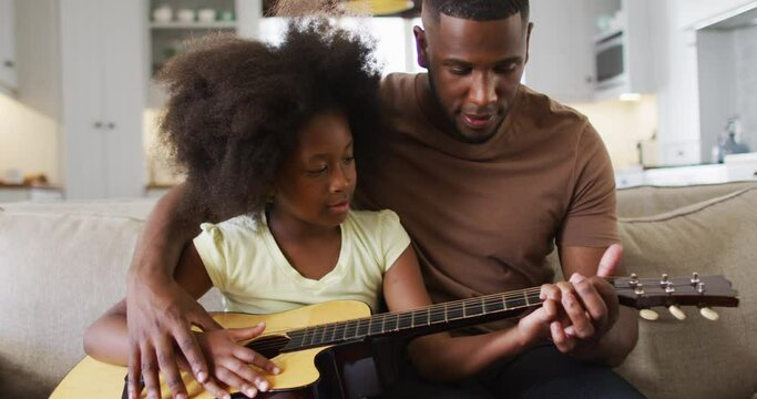 African american father and his daughter sitting on couch playing guitar together