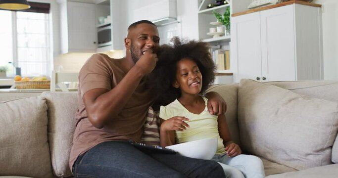 African american daughter and her father on couch eating popcorn and laughing at tv