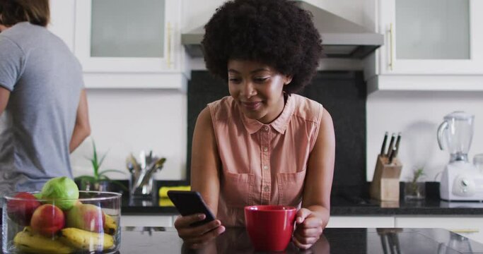 African american woman holding coffee cup using smartphone in the kitchen at home