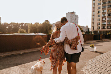Guy in white shirt and shorts is hugging his girlfriend in polka dot outfit. Couple walking their white labrador
