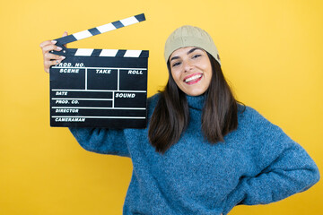 Young beautiful woman wearing blue casual sweater and wool hat holding clapperboard very happy...