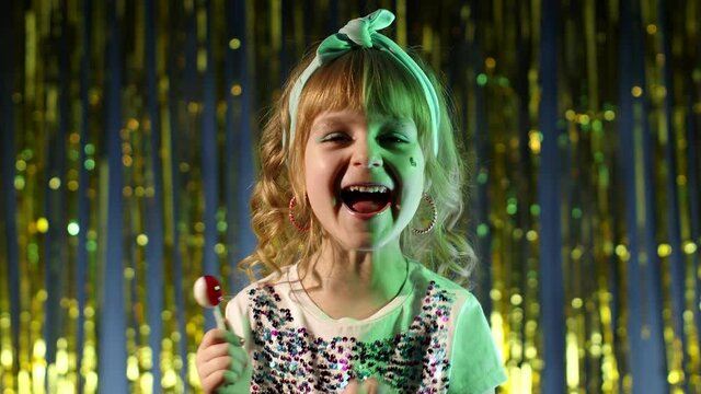 Trendy stylish girl with lollipop candy looking at camera smiling, screaming, shouting. Child kid in neon lights posing at disco club. Futuristic hipster teenager children making faces, fooling around