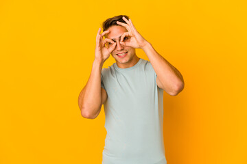 Young handsome caucasian man isolated showing okay sign over eyes