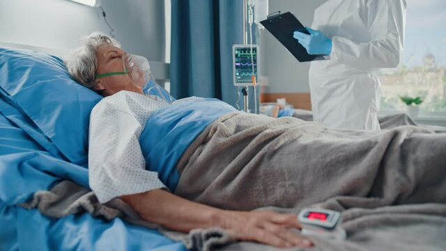 Hospital Coronavirus Emergency Department Ward: Doctor wearing Coverall Writes Down Vitals of a Senior Female Patient with Oxygen Mask Resting in Bed. Medics, Paramedics Doing everything to Save Lives
