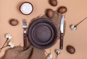 Ceramic brown plate onsustainable burlap cloth with Easter eggs, bowl with white cream sauce and cotton flowers on beige background. Zero waste holiday card. Eco-friendly clay dishes mockup