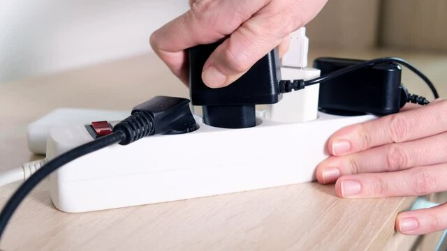 Hand plugging socket into electrical power strip overloaded with multiple electrical cords plugged in, many plugs plugged into electric power bar