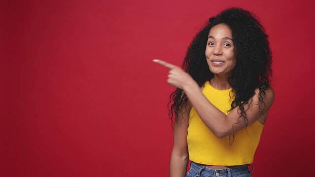 Laughing excited african american young woman in casual yellow tank top posing isolated on red background studio. People lifestyle concept. Pointing index finger aside showing thumbs up like gesture