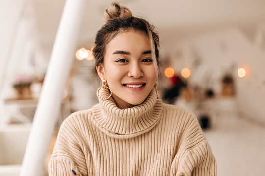 Lady in stylish beige oversize sweater looks into camera. Shot of girl with bun in massive earrings in bright room