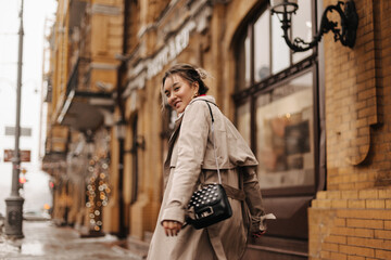 Fototapeta premium Young Asian girl in high spirits walks through city in stylish trench coat with small black bag